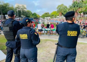 Três guardas civis municipais uniformizados patrulhando área pública com pessoas ao fundo