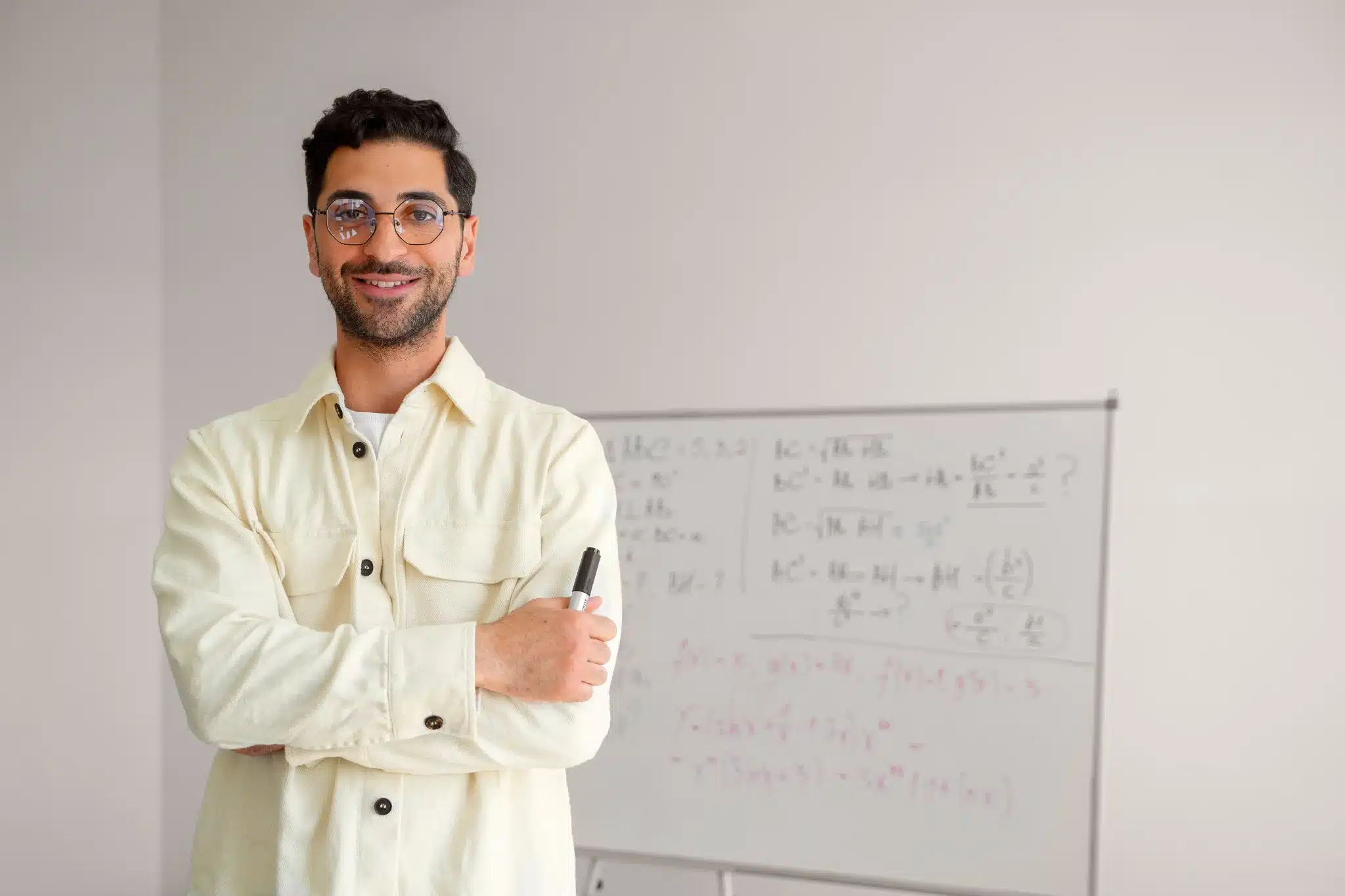 Professor sorridente com quadro branco e marcador Homem jovem usando óculos e camisa clara com marcador na mão, ao lado de quadro branco com equações matemáticas