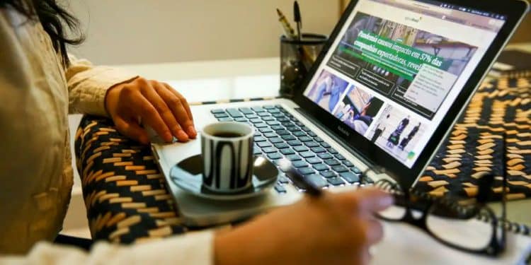 Mulher trabalhando remotamente em casa, digitando no laptop com xícara de café e caderno para ilustrar o conceito de teletrabalho.