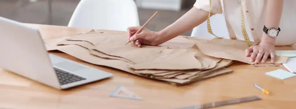 Criação de Moldes e Design Têxtil Detalhe de uma profissional de moda criando moldes de papel com lápis e régua, ao lado de um notebook.