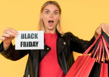 Mulher loira segurando cartão escrito Black Friday e várias sacolas vermelhas e pretas.