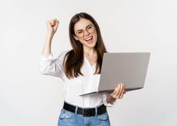 Mulher jovem e sorridente com óculos segurando um laptop cinza, fazendo gesto de comemoração com o punho.