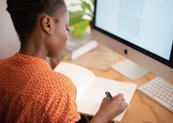 Jovem profissional independente trabalhando em home office, anotando ideias em caderno enquanto visualiza texto em monitor.