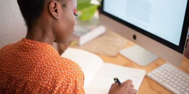 Mulher negra atenta escrevendo em caderno enquanto estuda curso online sobre compensação previdenciária em computador iMac.