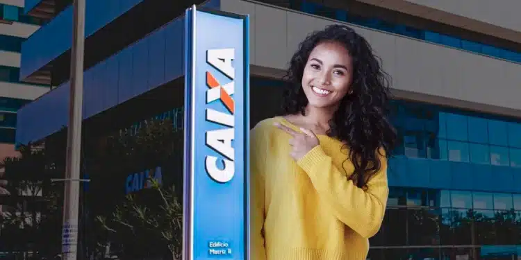 Mulher jovem de cabelo cacheado e suéter amarelo apontando para placa azul da Caixa Econômica Federal em frente a edifício empresarial