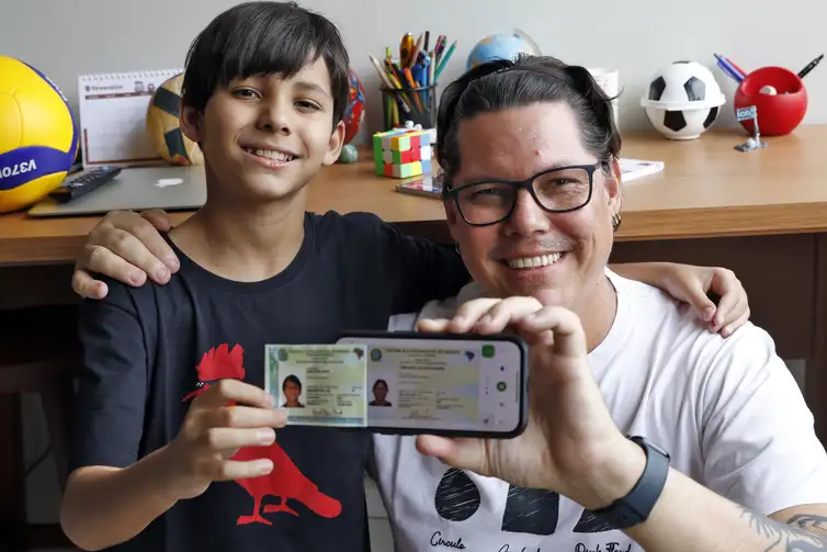 Pai e filho mostram documento digitalizado no celular Homem e menino sorridentes exibem documento de identidade digital em celular na frente de mesa com objetos