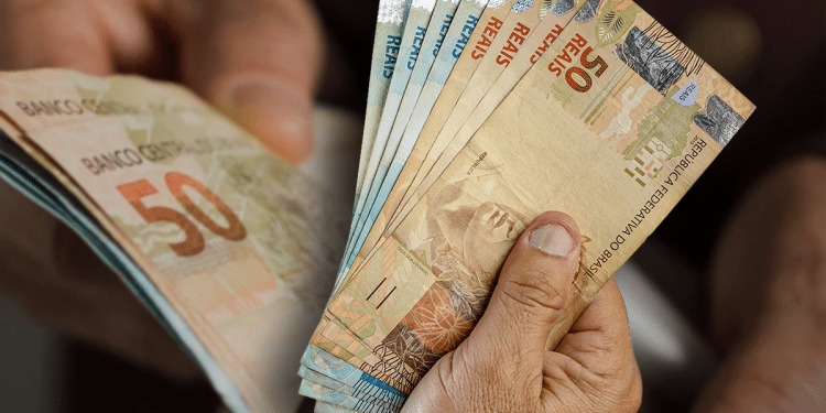 Mão segurando um leque de notas de cinquenta e cem reais, representando o dinheiro injetado na economia pelo décimo terceiro.
