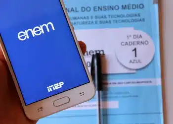 Tela de smartphone exibindo o aplicativo Enem e caderno azul do 1º dia do exame com caneta ao fundo