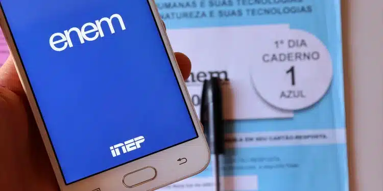 Tela de smartphone exibindo o aplicativo Enem e caderno azul do 1º dia do exame com caneta ao fundo