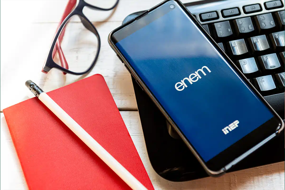 ENEM 2025: Organização para o Segundo Dia de Provas Telefone celular exibindo o logotipo "ENEM" e "INEP" em uma tela azul, ao lado de um teclado de computador, óculos de leitura e um bloco de notas com lápis, representando o estudo e a consulta de informações online para o Exame Nacional do Ensino Médio.