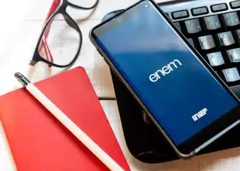 Aplicativo do ENEM em um smartphone sobre um teclado de notebook, ladeado por um bloco de notas vermelho e um lápis, simbolizando os estudos para o Exame Nacional do Ensino Médio e sua nova função como avaliação da qualidade do ensino médio a partir de 2026.