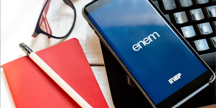 Aplicativo do ENEM em um smartphone sobre um teclado de notebook, ladeado por um bloco de notas vermelho e um lápis, simbolizando os estudos para o Exame Nacional do Ensino Médio e sua nova função como avaliação da qualidade do ensino médio a partir de 2026.