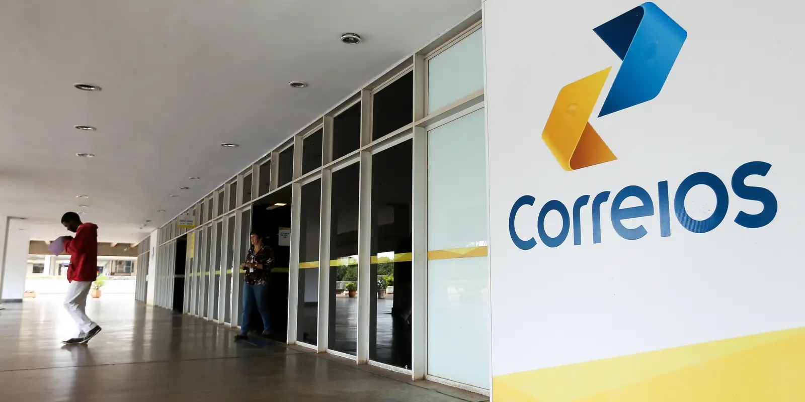 Fachada dos Correios mostra atendimento ao cliente Fachada da agência dos Correios com logo colorido e pessoas dentro e fora do prédio