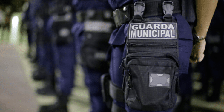 Uniforme da Guarda Municipal com bolsa preta e placa em destaque durante formação noturna
