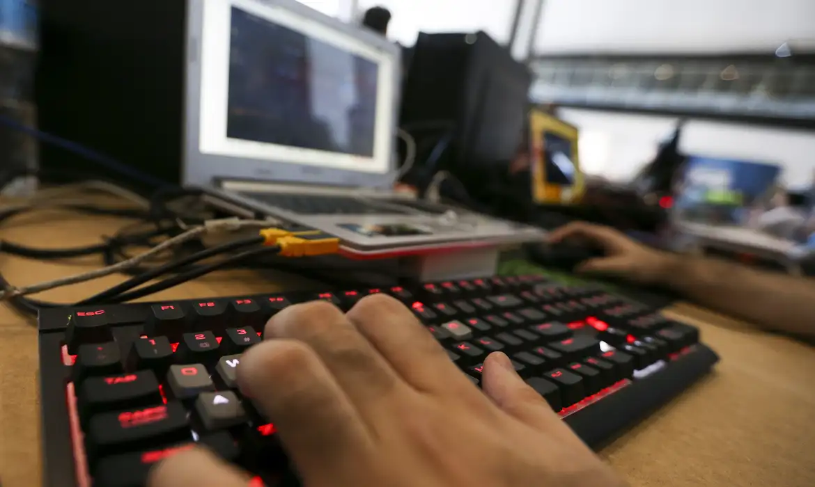 Pessoa digitando em teclado com laptop aberto Mão digitando em teclado iluminado vermelho com laptop e equipamentos eletrônicos ao fundo