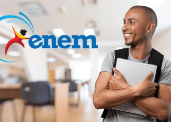 Jovem sorridente de camiseta cinza, carregando notebook, ao lado do logo do Enem em ambiente de sala de estudos.