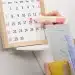 Mão marcando o dia 18 no calendário com marca-texto rosa segurando documentos e livro amarelo