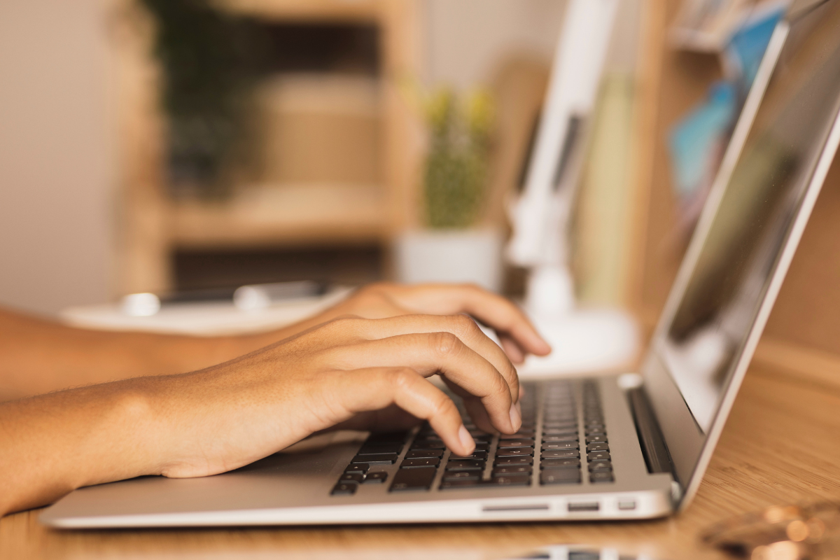 Inscrição Mãos femininas digitando em teclado de notebook prateado sobre mesa de madeira clara em escritório.