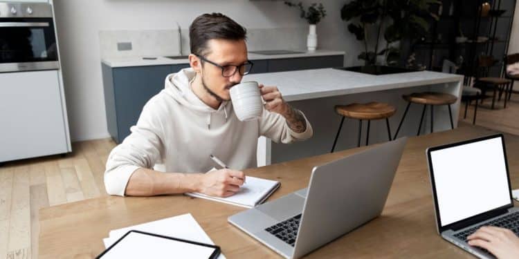 Homem jovem e focado trabalhando remotamente em uma cozinha moderna, usando notebook e tablet, enquanto bebe café e anota em um caderno.
