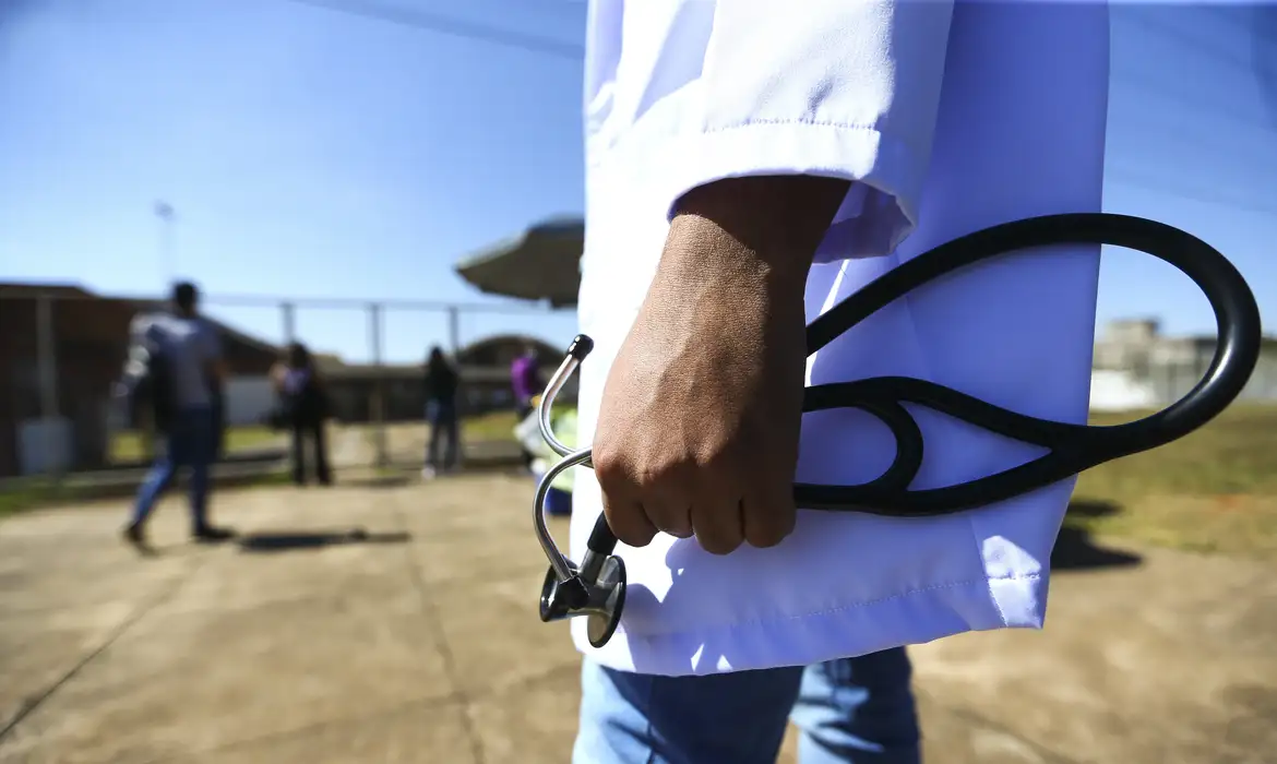 Médico segurando estetoscópio ao ar livre Médico de jaleco branco segurando um estetoscópio preto com pessoas ao fundo desfocadas em área externa