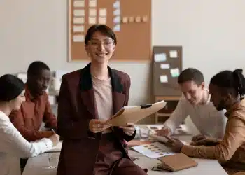 Mulher sorridente usando óculos e terno segurando pasta enquanto equipe diversa trabalha em mesa ao fundo