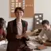 Mulher sorridente usando óculos e terno segurando pasta enquanto equipe diversa trabalha em mesa ao fundo