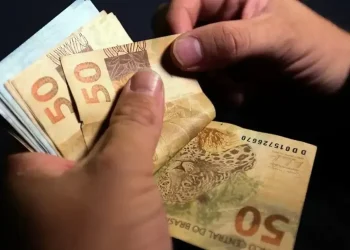 Mãos segurando múltiplas notas de cinquenta reais representando pagamento de R$ 200 ou R$ 1.000 do Pé-de-Meia