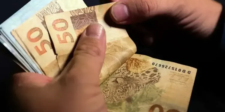 Mãos segurando múltiplas notas de cinquenta reais representando pagamento de R$ 200 ou R$ 1.000 do Pé-de-Meia