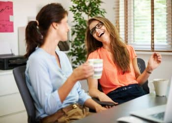 Duas mulheres conversando e sorrindo enquanto tomam café em ambiente de escritório iluminado