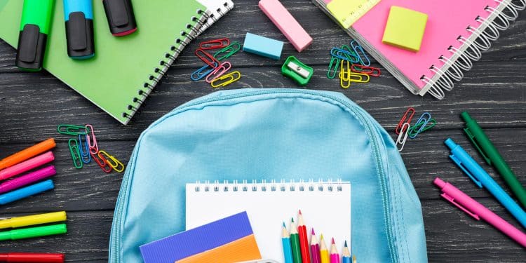 Mochila azul clara com caderno, lápis de cor, canetas, marca-texto e clipes coloridos sobre mesa preta.