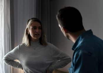 Mulher com blusa cinza conversando seriamente com homem de camiseta azul dentro de casa.