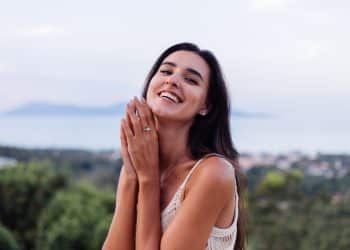 Mulher jovem sorridente com cabelo escuro, usando vestido branco e anel de prata nas mãos ao ar livre Legenda: Jovem feliz exibindo seu anel de prata em ambiente ao ar livre com paisagem desfocada ao fundo.