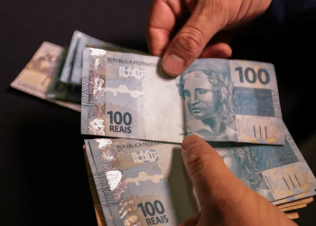 Mãos segurando e contando várias cédulas de cem reais em fundo escuro