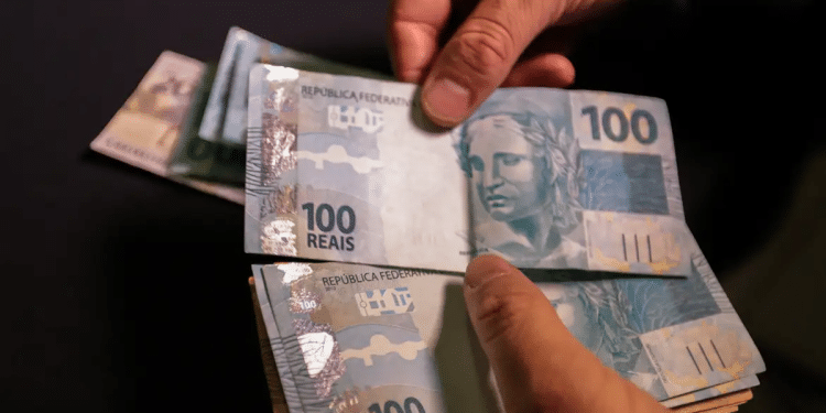 Mãos segurando e contando várias cédulas de cem reais em fundo escuro