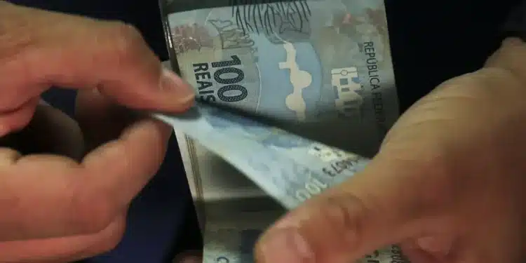 Mãos contando cédulas de cem reais representando o pagamento de mil reais do incentivo Pé-de-Meia.