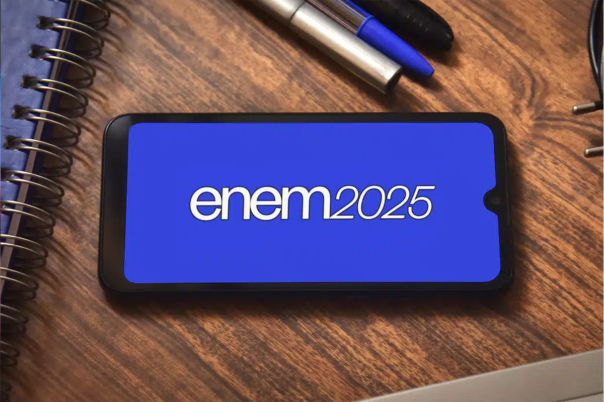 Cancelamento parcial do Enem 2025. Smartphone mostrando a tela do Enem 2025 sobre mesa com caderno e canetas, ilustrando alerta de cancelamento parcial.