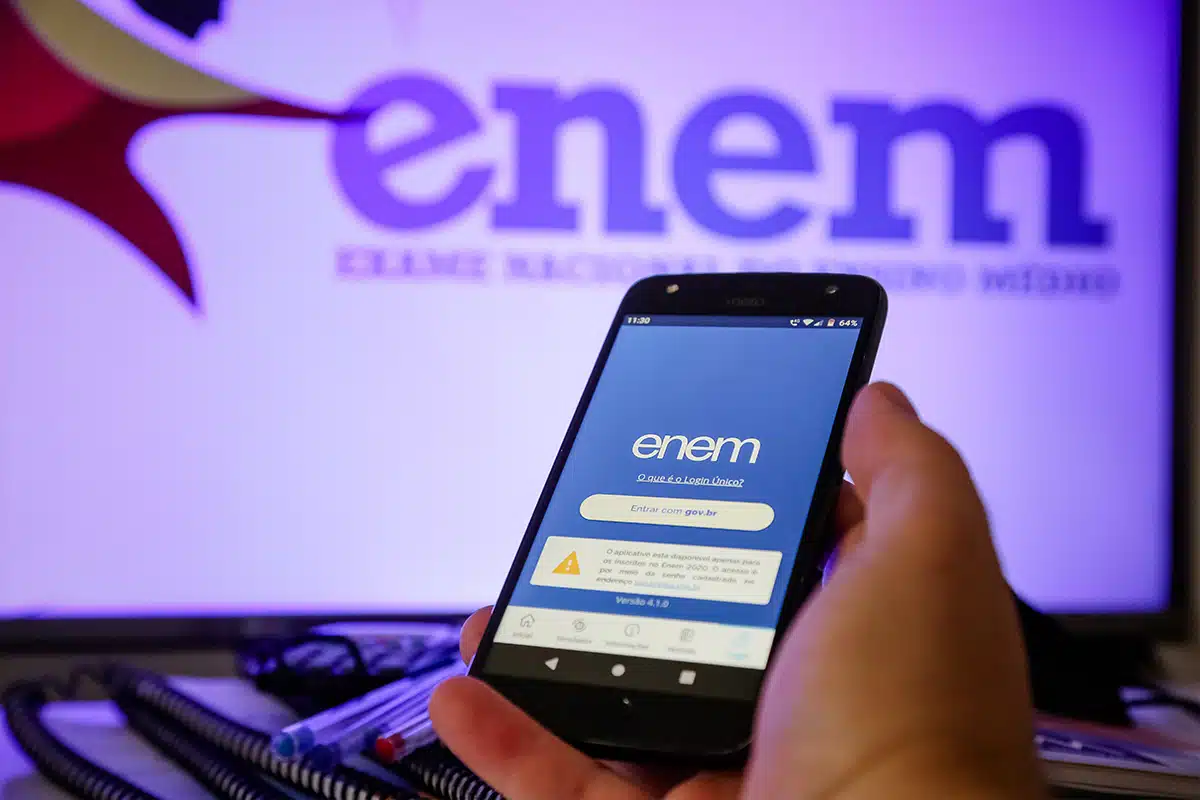 Enem 2025 Pessoa acessando o portal do Inep para ver o resultado do Enem 2025 em um celular, com o logo do Enem ao fundo na tela do computador.