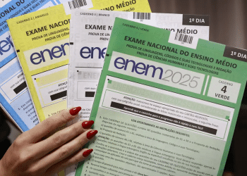 Mão segurando provas do ENEM 2025 com cadernos azul, amarelo, branco e verde.