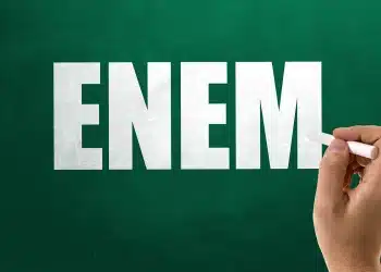 Mão escrevendo a palavra ENEM em um quadro verde, simbolizando debate sobre suspeita de vazamento da prova.