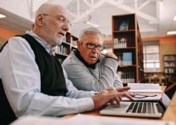 Dois homens maduros trabalhando juntos em frente ao computador em ambiente de estudo, em busca de novas profissões.