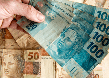 Mão segurando quatro notas de 100 reais sobre notas de 50 reais ao fundo.