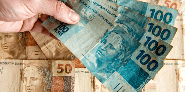 Mão segurando quatro notas de 100 reais sobre notas de 50 reais ao fundo.