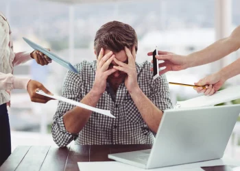 Homem estressado e sobrecarregado no trabalho, com múltiplas tarefas sendo apresentadas ao seu redor, simbolizando o esgotamento profissional (burnout).