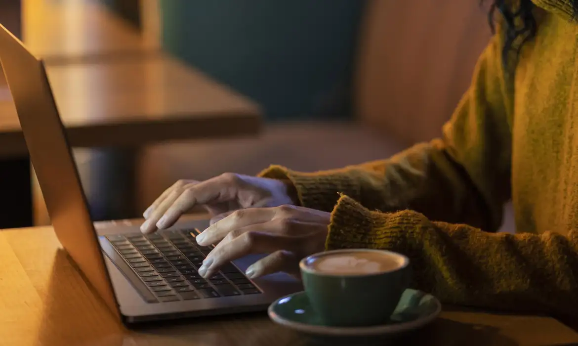 Pessoa digitando no laptop com café ao lado Mãos digitando no teclado de um laptop ao lado de uma xícara de café em ambiente aconchegante