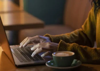 Mãos digitando em laptop sobre mesa de madeira com xícara de café ao lado à noite
