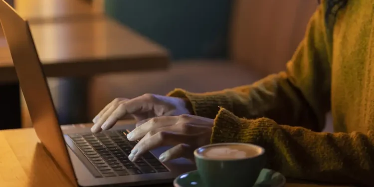Mãos digitando em laptop sobre mesa de madeira com xícara de café ao lado à noite