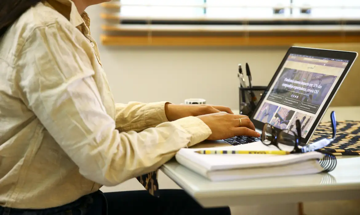 Pessoa digitando no notebook em ambiente de trabalho Mulher usando camisa bege digitando em notebook sobre mesa com caderno, lápis e óculos