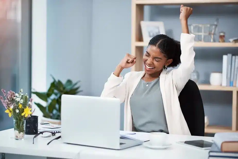 Estratégias para ser feliz no trabalho. Mulher comemorando com braços erguidos em frente ao notebook, representando a satisfação profissional e o conceito de job crafting no escritório.