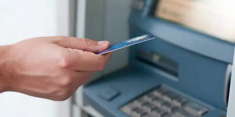 Pessoa utilizando um caixa eletrônico de banco com cartão na mão, em frente à tela do terminal.