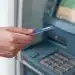 Pessoa utilizando um caixa eletrônico de banco com cartão na mão, em frente à tela do terminal.
